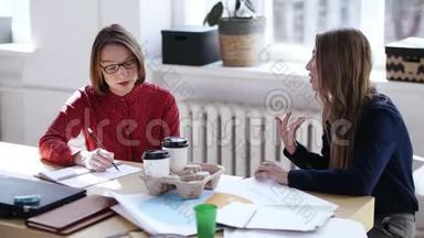 两位漂亮的经理妇女在休息时在<strong>现代办公桌</strong>上和咖啡聊天。 女老板听同事讲话